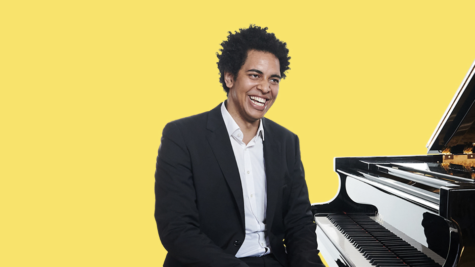 A male student with black hair, wearing a white shirt and a black suit, sitting at a piano, smiling at the camera, with a yellow background behind him.